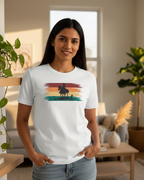 Woman wearing a white t-shirt with a graphic design of a person on horseback during sunset, standing in a living room.