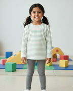 Young girl in a striped shirt and gray pants standing in front of colorful building blocks.