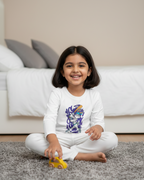 Young girl sitting on a carpeted floor holding a toy car, wearing a white shirt with a colorful design.
