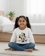 Young girl sitting on a carpeted floor wearing a white sweatshirt with a cartoon character, in a room with a bed and plant in the background.