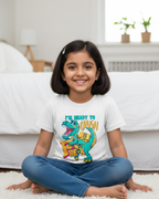 Young girl sitting on a carpeted floor wearing a white t-shirt with a colorful dinosaur design.