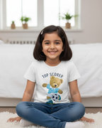 Young girl sitting on a carpeted floor wearing a white t-shirt with a teddy bear design and text.