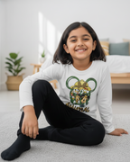 Young girl sitting on the floor wearing a white long-sleeve shirt with a colorful graphic design.