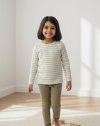 Young girl wearing a striped shirt and beige pants standing in a bright room with white walls and wooden floor.
