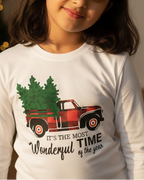 Young girl wearing a white long-sleeve shirt with a Christmas design, standing in a festive room with a decorated tree and presents.