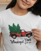 Young girl wearing a white t-shirt with a Christmas design, standing in a room with a wooden shelf and plant.