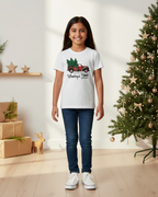 Young girl wearing a white t-shirt with a Christmas design, standing in front of a decorated Christmas tree.